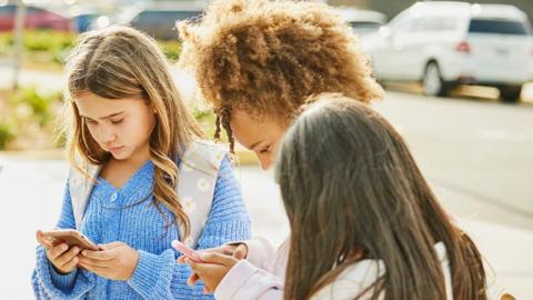 The image shows children standing in a circle looking at their phones outside a school.