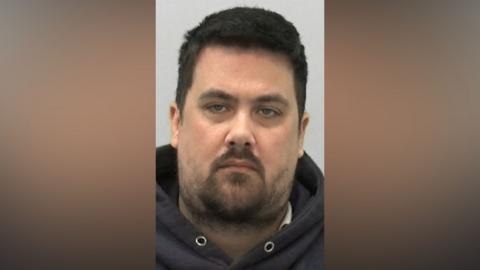 Mugshot of Scott McKenzie. He has a large head with a wispy black beard and short thick black hair.