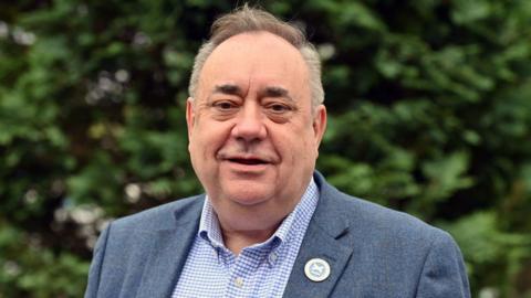 Alex Salmond smiling in medium close up. He is weating a blue tweed jacket, open-necked button down white and blue checked shirt and an Alba party badge