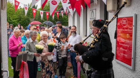 Many people, young and old, gathered outside Kilchoan Post Office to celebrate its reopening at a new location. A piper is stood outside the door in traditional dress, playing his bag pipes.