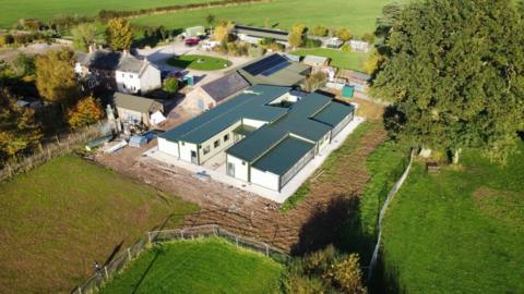 An aerial view of the new kennels. It is a large, white one-storey building with flat blue ceiling. It is surrounded by several other buildings and green fields.