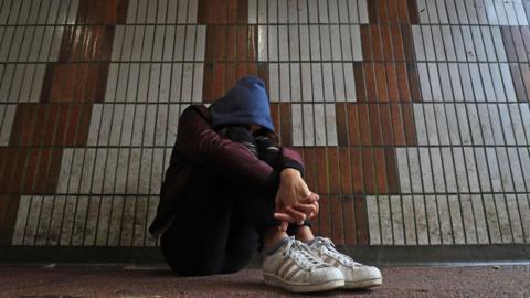 Illustrative photograph, posed by a model, of an anonymous young girl sitting with her head lowered and her hands clasped around her knees.