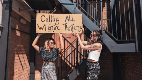 Two women hold up a homemade cardboard sign reading 'calling all Wiltshire thrifters', while standing in front of an outdoor metal staircase.