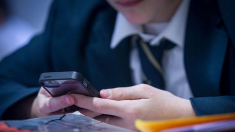 A school pupil wearing a shirt, tie and blazer holding a mobile phone in a classroom.