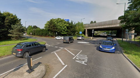 The junction at Holyport Road and the A308 Windsor Road, where there is a mini roundabout with three exits. There is a concrete motorway bridge over the road beyond one of the exits.