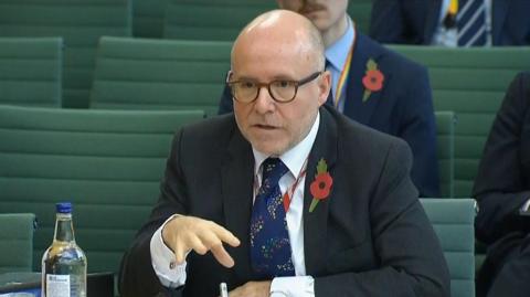 Lord Hermer speaking to Parliament's Joint Committee on the National Security Strategy. He is wearing a suit, red poppy and glasses. Behind him a rows of green chairs and other people watching the committee.
