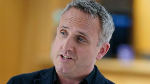 The leader of the Scottish Liberal democrats, Alex Cole-Hamilton smiles as he look away from the camera. He has grey hair and blue eyes. He appears to be in the Scottish parliaments building but the background is blurred.