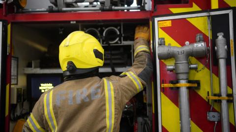 The backs of three firefighters wearing breathing equipment and helmets. A fire station and other firefighters are out of focus in the background.