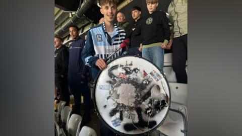 Cory stands next to his damaged drum. The skin is ripped, with the remnants showing the stickers fans have affixed to it over the years. Cory is wearing a Coventry City hooded top with 'Hummel' on the chest.
