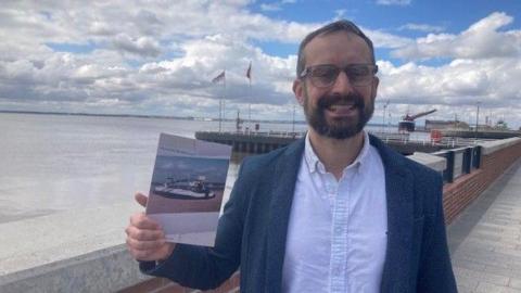 A man with a beard and glasses stands holding a book with a photo of a hovercraft on the front. He is wearing a white shirt and a blue blazer. He is by the harbour wall, with the river Humber in the background. 