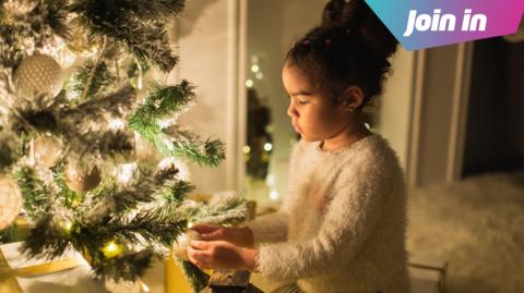 A girl with brown curly hair tied up in a bun wears a white fluffy jumper and hangs a decoration on a Christmas tree with white glowing lights on it