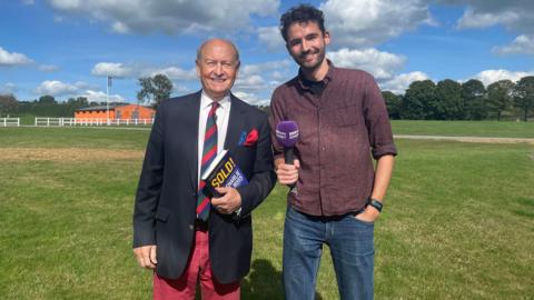 Charlie Ross, from Bargain Hunt, with Charlie Taylor from 鶹Լ Radio Somerset. Charlie Ross is wearing a suit jacket with red trousers and a striped tie. He is holding a copy of his autobiography, "Sold!". Charlie Taylor is standing next to him, holding a purple 鶹Լ Radio Somerset microphone. Both of them are looking at the camera and smiling. 