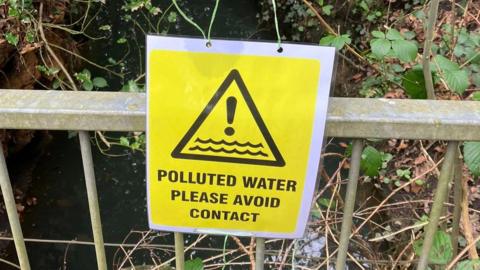 A yellow warning sign with the words POLLUTED WATER PLEASE AVOID CONTACT written in black. it is hung on grey metal railings