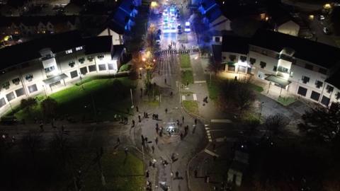 The image shows a residential area from above with lots of young people dressed in black. There is a line of police up the road with emergency service vehicles flashing blue lights behind them.