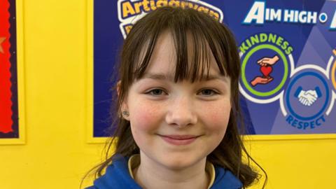 Alesha is wearing a blue hoodie and a yellow top underneath. She has dark hair with a fringe, freckles and is smiling.