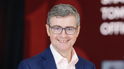 Brendan Mooney is wearing a blue jacket and light pink open-necked shirt. He has grey hair and is wearing dark-rimmed glasses and is smiling