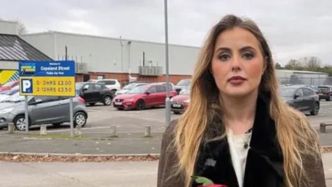 Miss Hudson, who is a young blonde woman, stands outside the Excel carpark in question near a sign showing charges, wearing a brown coat 