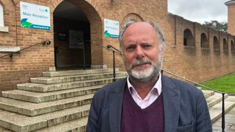 Julian Brazil is wearing a light pink shirt and a maroon V-neck sweater under a dark blue suit jacket and is standing in front of steps at the main entrance to the red brick-fronted County Hall in Exeter.