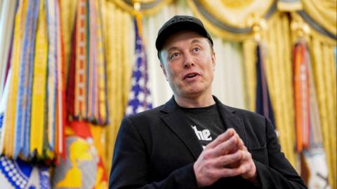 Elon Musk, wearing black, with hands together, fingertips touching, and a baseball cap.