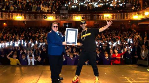 A packed theatre audience fills a large, ornate hall with glowing lights held aloft throughout the crowd. On stage, two individuals stand at the front, holding up a framed certificate together. One person is dressed in a dark suit, while the other wears a black T‑shirt, gold accessories and brightly coloured shoes. Rows of people, including many children in school uniforms, cheer and raise their hands from the stalls and the balcony above. Warm, golden lighting illuminates the interior, highlighting the decorative railings and wood‑panelled walls surrounding the scene.