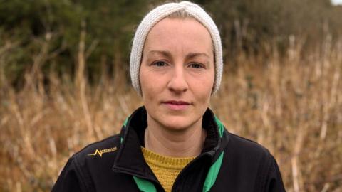 a woman in knitted clothing looking at the camera - behind her is a lot of tall grass