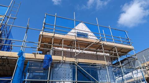 Stock photo shows the shell of a house with scaffolding around it against a blue cloudy sky, illustrating the building of new housing.