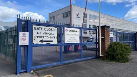An entrance to the Vauxhall factory in Luton. There is a blue gate with a Vauxhall sign on it and another sign saying "Gate Closed". In the background is a large building with another Vauxhall sign on it.