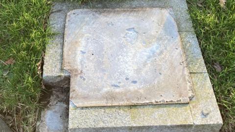 A square stone plinth in the Gardens of Peace. A corner chunk of stone is missing from the plinth and the memorial plaque which usually rests on top has gone. The plinth is surrounded by grass.