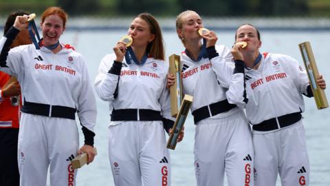 Four rowers wearing white tracksuits. All of them have a gold medal around their necks, holding it up or kissing it in victory