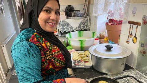 A smiling woman stood in front of a cooker. She is wearing a turquoise dress with a colourful pattern on the front and a black head scarf. There is a large pan in front of her with other dishes containing food on a counter top. 