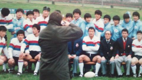 A wax jacket-wearing photographer takes a photo of the South Korean rugby team sat in rows, many wearing white shirts with red and blue hoops on them, and others in blue track suits, while mangers wore blue blazers.