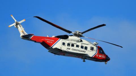 A red and white coastguard helicopter flies through a bright blue sky