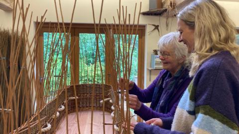 A half built willow coffin is being woven by felicity who is wearing a purple jumper and glasses and Caz who is standing next to her in a mixed purple, light blue and cream jumper