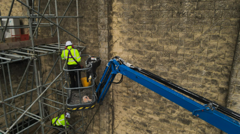 A contractor in a cherry picker and one on scaffolding working to install new heating