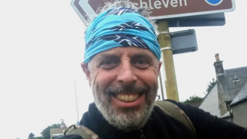 A smiling man with a grey beard and blue head scarf standing outside