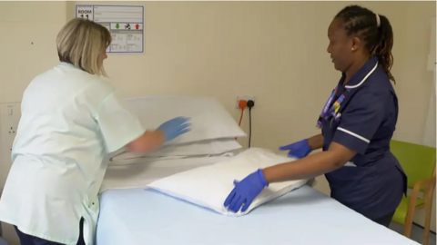 Two nurses, one in a light uniform and another with a blue uniform prepare a hospital bed with a light wall in the background and three pillows on the bed.