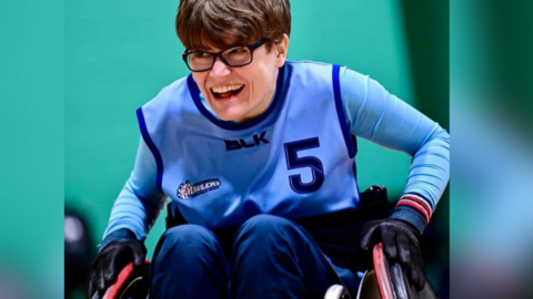 Phoebe Gibbons in her blue kit sat in a wheelchair rugby chair. She is also wearing glasses and smiling. She's wearing gloves and her hands are resting on the wheels. 