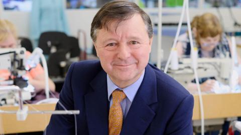 Christopher Nieper is wearing a blue suit and orange tie, he is smiling at the camera and there are sewing machines in the background. 