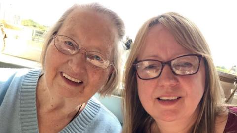 Norma Graham is on the left. She is an elderly lady wearing a blue jumper and has glasses and grey hair. Carole Parkin is next to her, she is younger and has brown hair and glasses 