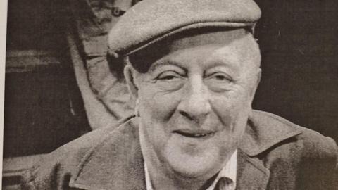 A black and white photo of a man smiling, wearing a hat and jacket, with white shirt underneath