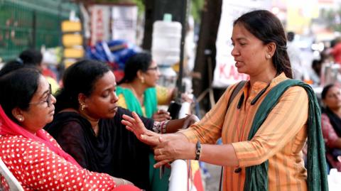 A protest coordinator Saboora Arifa, seen in a yellow kurta and green dupatta, standing across two protesting volunteers - one in red, one in black. 