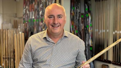 A middle-aged man with a patterned button shirt holds a snooker cue aloft.