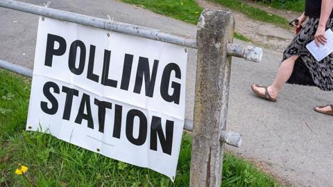 A polling station in Shropshire. There is a metal gate with a paper sign that says POLLING STATION. Someone can be seen walking past with a ballot paper in the background 