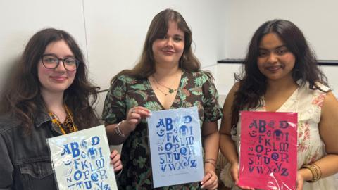 Three young women standing next to each other in front of a white background. They are holding posters with the alphabet printed on them in different fonts.