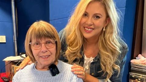 Theresa Carr is sitting down in a knitted jumper, wearing glasses, with brown hair. Louise Pentland, who has blonde hair and a blue denim jacket, leans over her with her hands on her shoulders. They are in a BBC radio studio with blue painted walls.