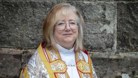 The Very Revd Manon Ceridwen James. She has short blonde hair and glasses and smiles at the camera. She wears gold trim robes with a gold pattern on. 