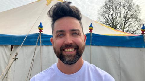 A man in a white t-shirt smiles in front of a tent. He has dark hair, blue eyes and a short beard. In the distance, a tree can be seen over his left shoulder.