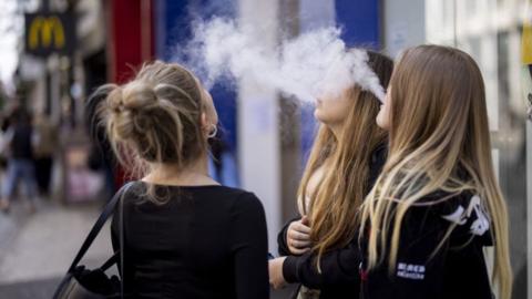 Young girls vaping. They are blowing out smoke and have their faces obscured or turned away from the camera