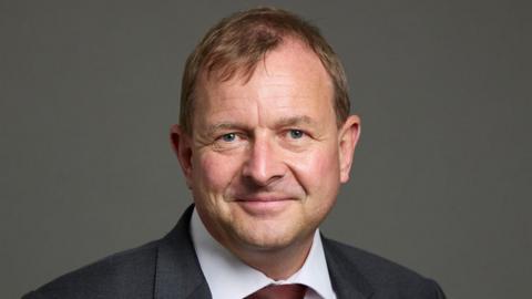 Lord Fuller in a dark jacket, a white shirt and red tie looks straight at the camera 