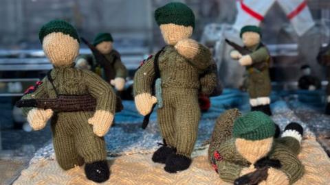 Five knitted British soldiers in The Longest Yarn exhibition. The knitted soldiers are wearing light green uniforms and dark green berets. They are holding black knitted rifles and are standing one a light cream knitted base to depict the sand of Normandy's beaches. One of the soldiers is lying down and aiming down his rifle..